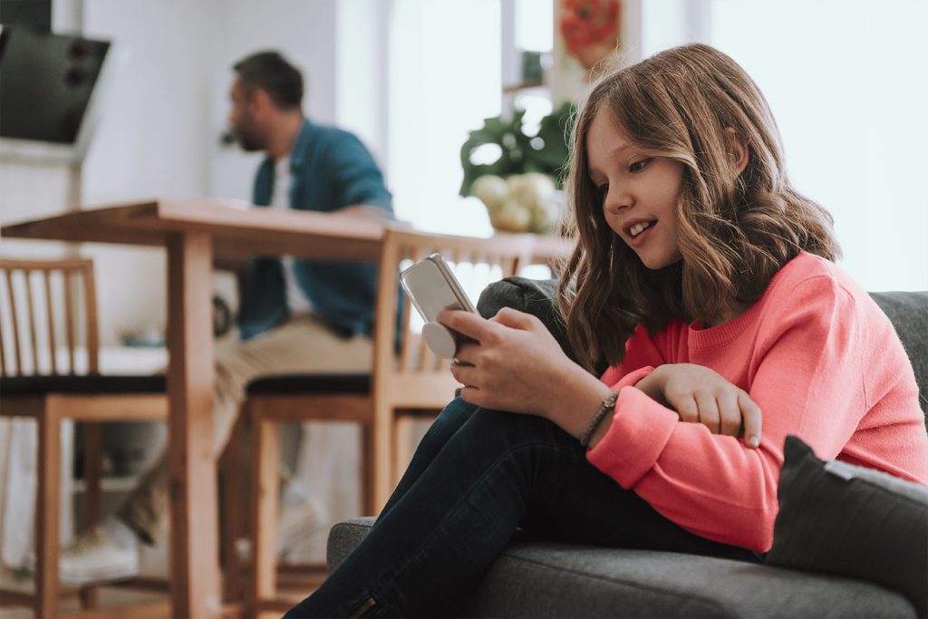 Young Teen girl playing on her phone in the living room with her dad in the background at the kitchen table